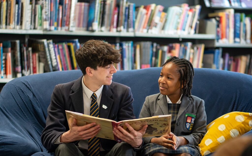 A senior student and a younger student sit together on a blue sofa in a school library, sharing a book and smiling at each other. The scene highlights mentorship and a supportive learning environment, with bookshelves in the background.