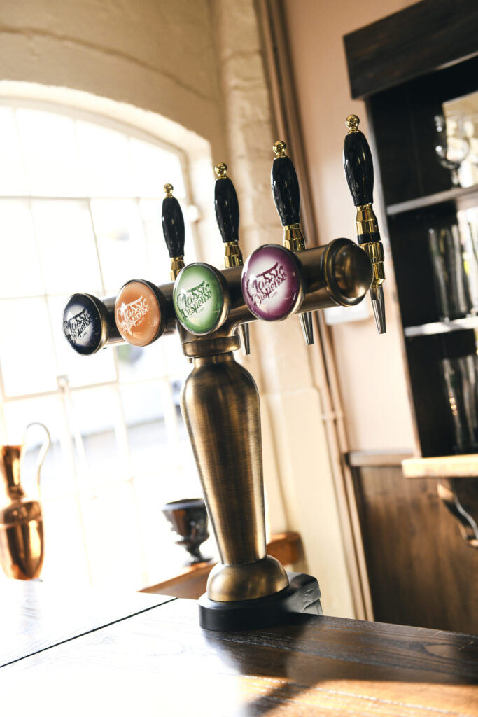 Four vintage-style beer taps with colourful branding, including black, orange, green, and purple handles, displayed on a polished brass tower in a warmly lit bar setting with large windows in the background.